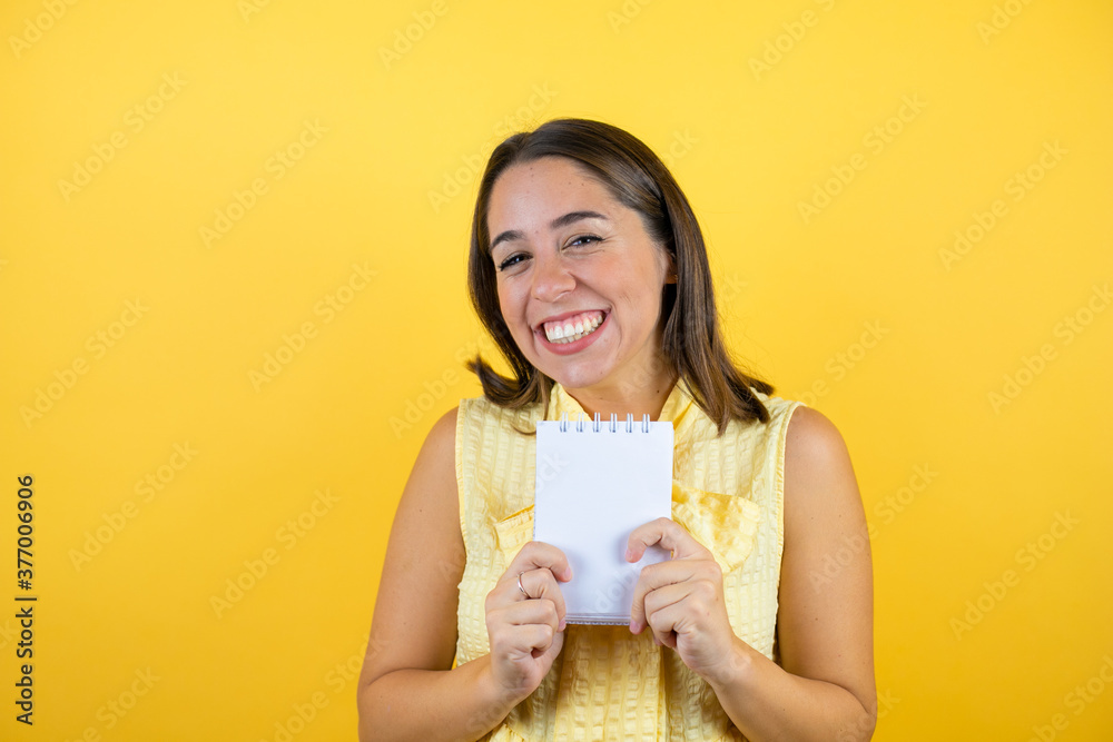 Young beautiful woman over isolated yellow background smiling and showing blank notebook in her hand Young beautiful woman over isolated yellow background smiling and showing blank notebook in her hand