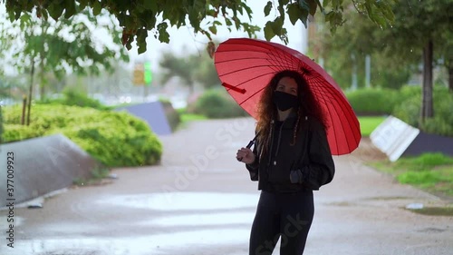 Slow motion video of a spanish woman wearing a mask on a park in Palma (Spain) walking to camera with a beautiful red umbrella while it’s raining due to a cold front at the arribal of autumn to Spain photo