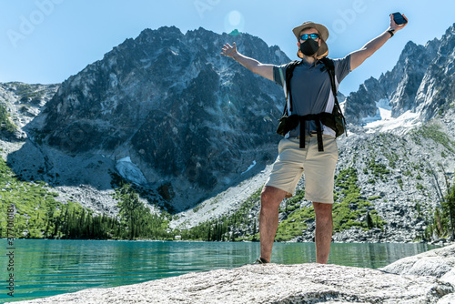 Hiker wearing protective face mask in the mountains with arms raised by lake.