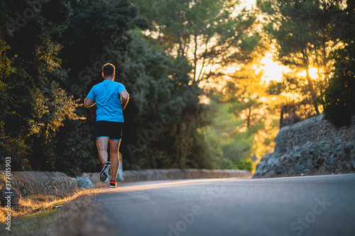 Young man wearing blue shirt and black shorts runs on an idyllic road during sunset surrounded by nature in Mallorca (Spain) © Daniel