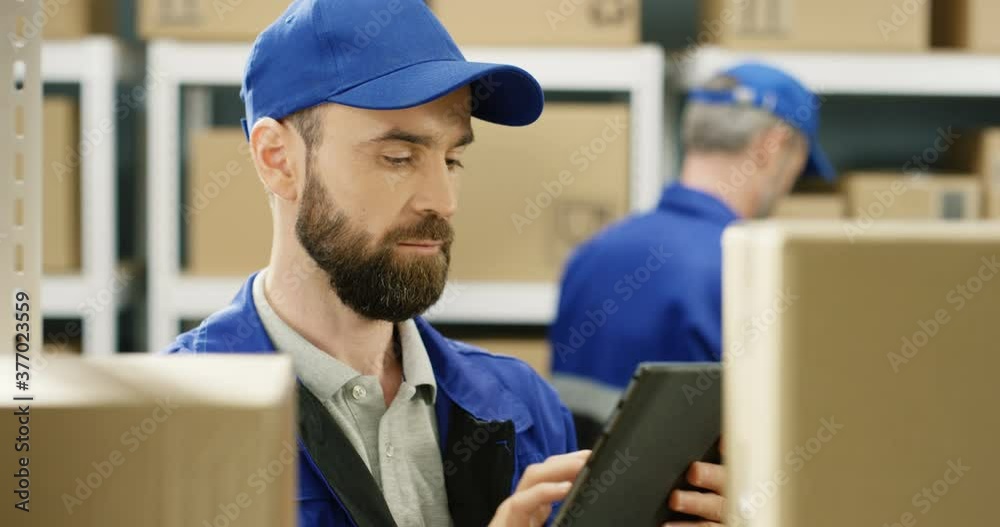 Close up of handsome Caucasian postman in blue uniform and cap standing between shelves with carton boxes in postal office storage. Man with tablet device searching for parcel and registering mails.