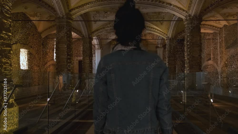 Adult woman walks inside Evora chapel of bones (Capela dos Ossos) on walls and pillars decorated, arranged and covered with human skulls and remains in cement, Portugal, static close up behind slowmo