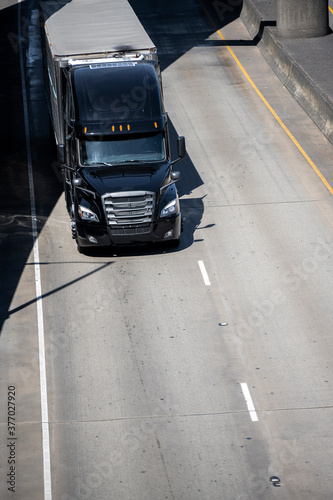 Stylish black big rig semi truck with semi trailer running under the bridge on the urban highway road