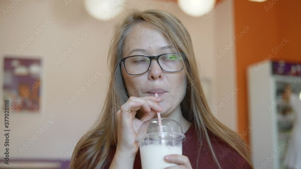 Beautiful blonde girl in glasses drinking milkshake from a plastic cup with straw sitting at cafe, close up Beautiful blonde girl in glasses drinking milkshake from a plastic cup with straw sitting at cafe, close up