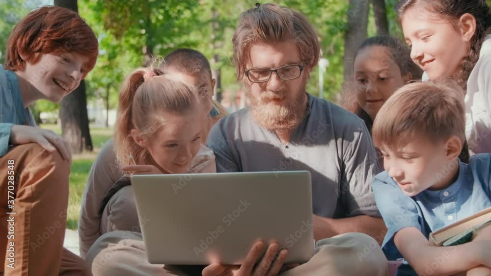 Sequence of shots of male teacher with laptop and curious schoolchildren gathered around him looking at laptop while sitting on blankets in park on summer day Sequence of shots of male teacher with laptop and curious schoolchildren gathered around him looking at laptop while sitting on blankets in park on summer day