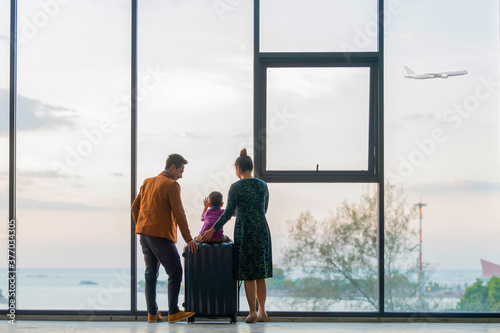 Happy asian family with suitcases in the airport. Little boy sitting on suitcase while Father and Mother standing near window at the airport and watching plane before departure on vacation.