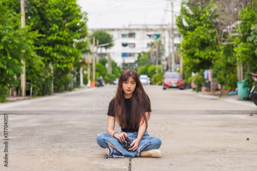 Close up portrait young Asian woman in a black T-shirt sitting on the floor holding a camera looking at the camera at the village road.