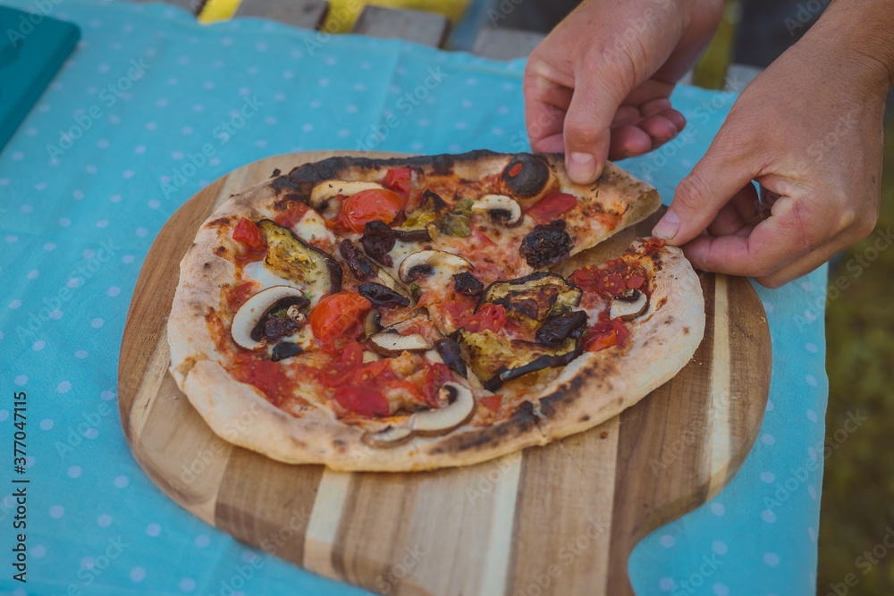 Home made pizza freshly baked and served on a wooden plate on a rustical table in a garden. Beautiful setting for a pizza, hands of a person are seen taking a slice.