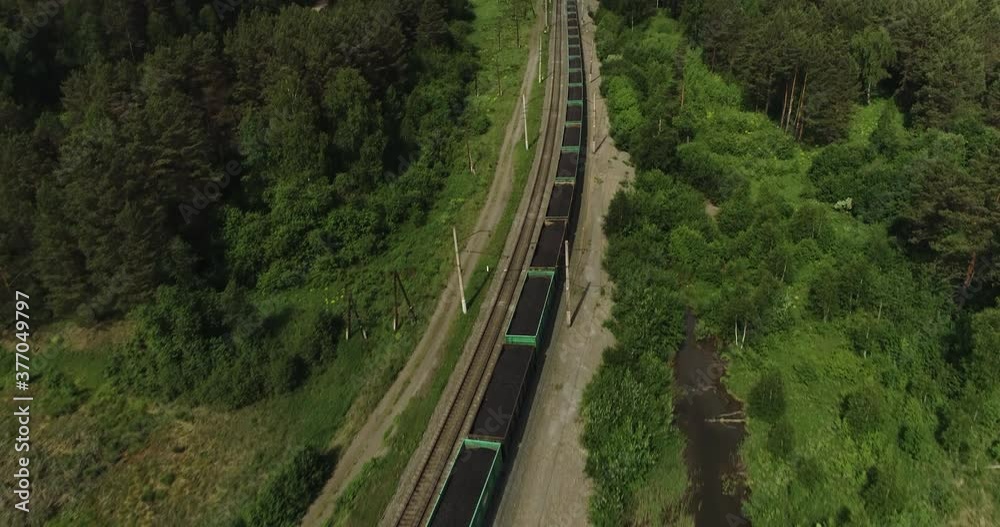 Freight long train carries with coal carriages an electric locomotive by two-sided Trans Siberian railways under the rock and near mountains river / Aerial drone wide view at summer sunny day Freight long train carries with coal carriages an electric locomotive by two-sided Trans Siberian railways under the rock and near mountains river / Aerial drone wide view at summer sunny day