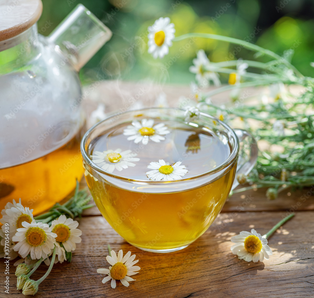 Herbal chamomile tea and chamomile flowers near teapot and tea glass on wooden table. Countryside background. Herbal chamomile tea and chamomile flowers near teapot and tea glass on wooden table. Countryside background.