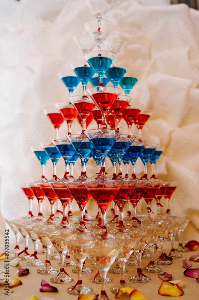 Naklejka premium Pyramid of glasses with Martini cocktail for a festive reception at the wedding on the table. Cosmopolitan with cherry, red, blue cocktail.