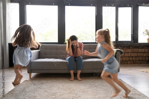 Young tired single mother suffers from headache closed eyes touch forehead sitting on couch while her daughters running around her and shouting  female babysitter feels exhausted by noisy kids