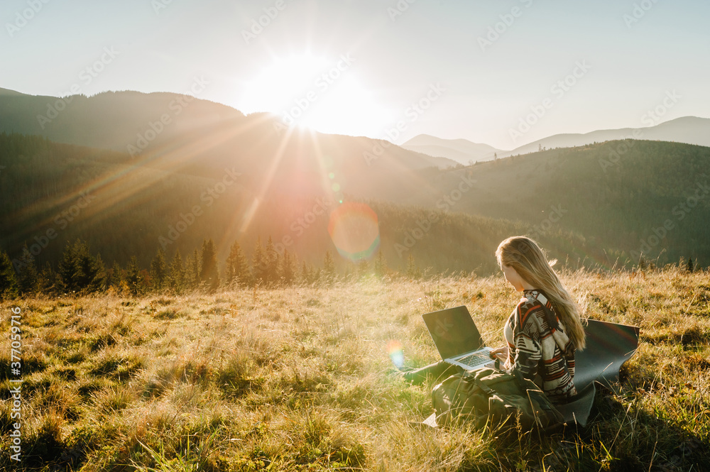 Woman working outdoors with laptop sitting in mountains. Concept of remote work or freelancer lifestyle. Cellular network broadband coverage. internet 5G. Hiker tourist enjoying valley view sunset. - obrazy, fototapety, plakaty 