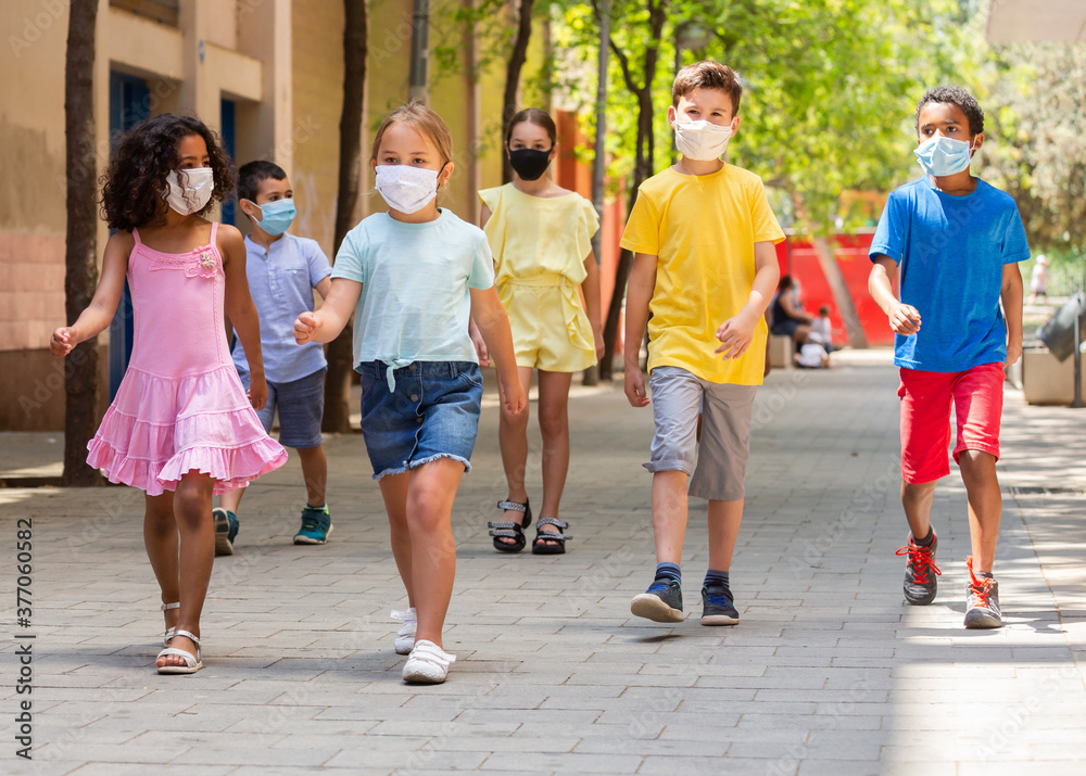 School children in protective medical masks walk along the street of a summer city
