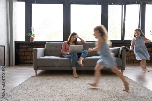 Young single mother working on laptop in loft sitting on couch while her daughters running around her and shouting