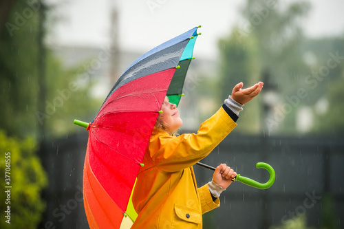 Happy girl portrait with bright rainbow umbrella. Children under the rain. Kid wearing yellow waterproof coat and boots holding colorful umbrella. Rainy weather. Child is catching drops in sunny day