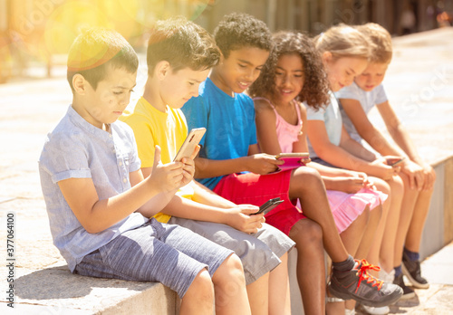 Group of positive children with mobile devices sitting at urban street
