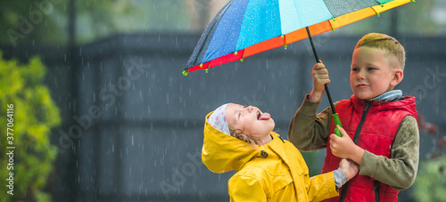 Little girl and boy with colorful umbrella playing in the rain. Kids play outdoor by rainy weather in fall. Autumn fun for children. Toddler kid in raincoat and boots walk in the garden