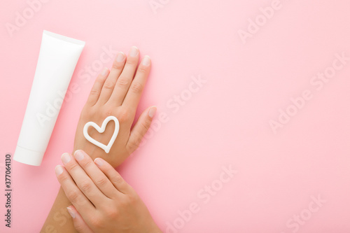 Heart shape created from cream on young woman hand. White tube. Care about clean and soft body skin. Closeup. Empty place for text on light pink table background. Pastel color. Top down view.