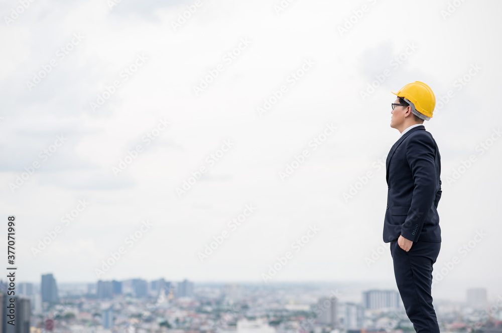 Young handsome asian civil engineer standing wearing helmet yellow hard hat in business suite on building urban cities background. Young handsome asian civil engineer standing wearing helmet yellow hard hat in business suite on building urban cities background.