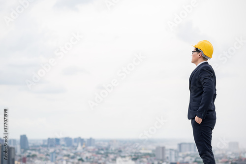 Young handsome asian civil engineer standing wearing helmet yellow hard hat in business suite on building urban cities background.