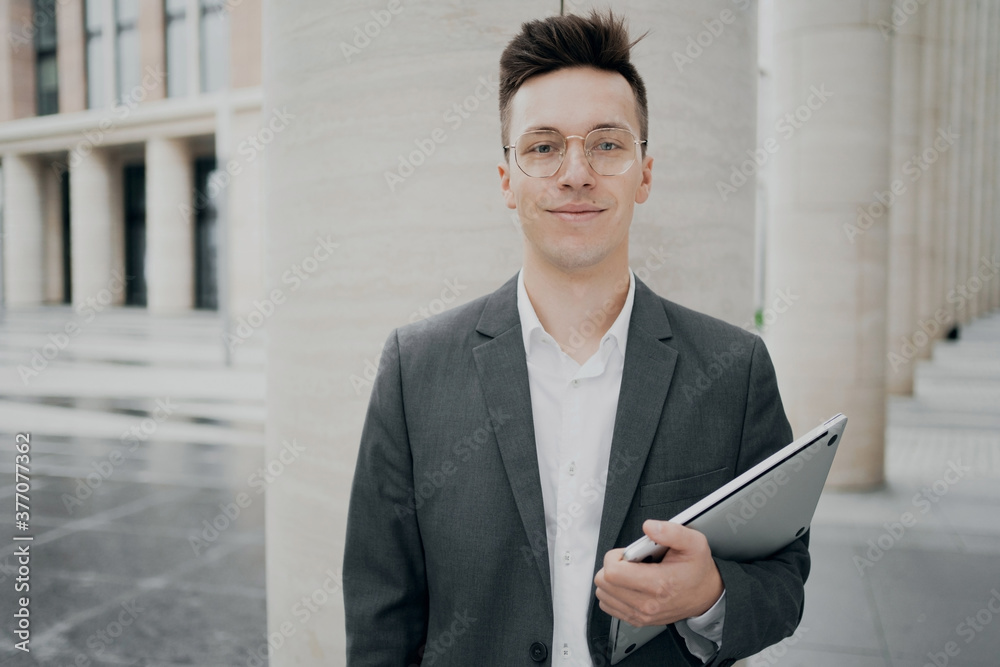 cheerful smart young man guy with glasses in a gray suit of European appearance, holding a laptop. gray business suit, standing near the business center in a hurry to work cheerful smart young man guy with glasses in a gray suit of European appearance, holding a laptop. gray business suit, standing near the business center in a hurry to work