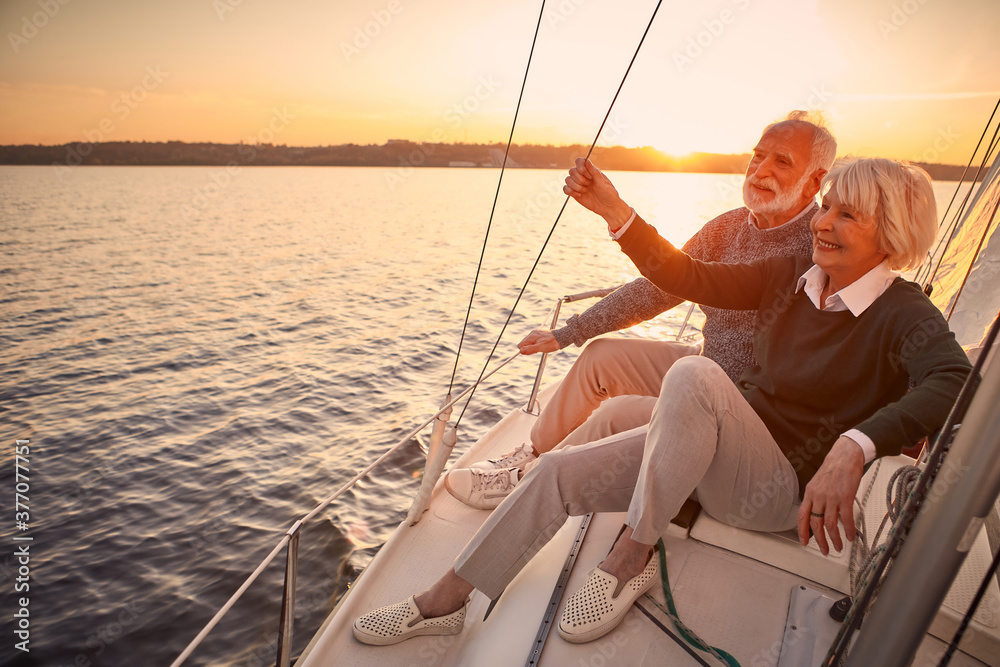 Enjoying luxury life. Beautiful happy senior couple in love relaxing on the side of sailboat or yacht deck floating in sea at sunset, looking at amazing evening view