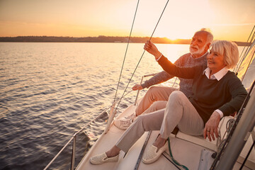 Enjoying luxury life. Beautiful happy senior couple in love relaxing on the side of sailboat or yacht deck floating in sea at sunset, looking at amazing evening view
