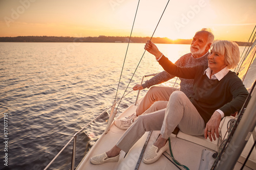 Enjoying luxury life. Beautiful happy senior couple in love relaxing on the side of sailboat or yacht deck floating in sea at sunset, looking at amazing evening view © Kostiantyn
