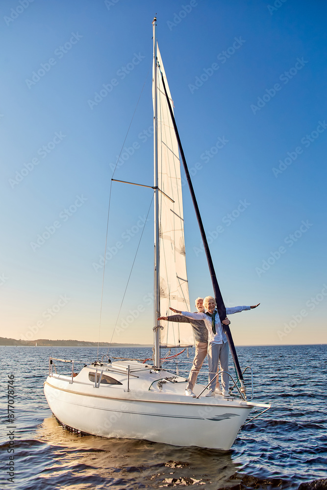 Happy senior couple stretching hands out against the sky, looking at the horizon and enjoying amazing view, standing on the side of sailboat or yacht deck floating in sea