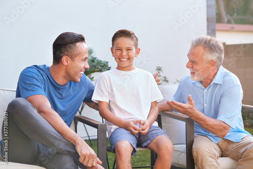 Male Multi-Generation Hispanic Family Sitting In Chairs Relaxing In Garden At Home Together 