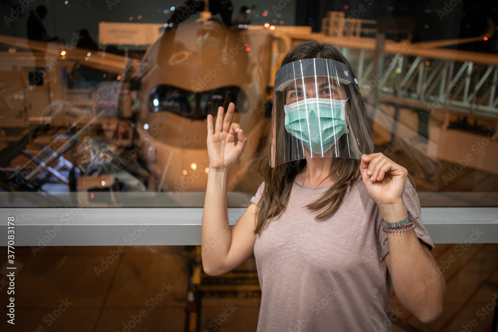 Female passenger ready to board. Woman wearing face mask and plastic face shield. Safety measures implemented by airlines in order to avoid risk of infection during Covid-19 pandemic. Airplane behind.