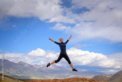 Young woman enjoying life and jumping against blue sky and mountains background. Happy female tourist jumps with hands up on top of rock on summer sunny day. Travel, freedom concept.