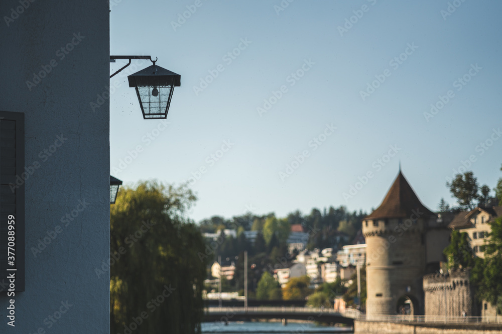 A lantern on a house wall by the Reuss river in Lucerne. In the background the powder tower of the Musegg Wall.
