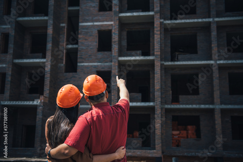 Back view a man and a woman in orange helmets stand with their arms around each other and look at a brick apartment building under construction. Investment in apartment, mortgage construction.