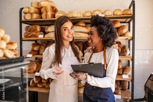Attractive women selling fresh pastry and loaves in bread section and smiling. Portrait of two beautiful young bakers at the bakery. Business owner talking to worker at a bakery