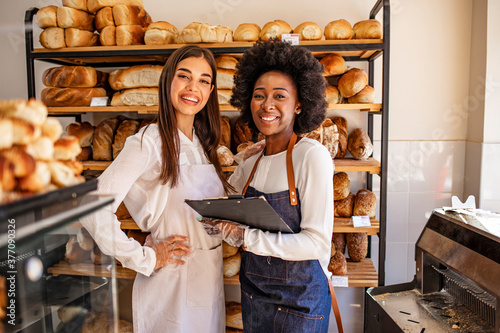 Two beautiful young bakers at the bakery. Shot of a young woman showing her colleague something on her clipboard while they stand in their bakery shop. Smiling young bakers at the bakery shop