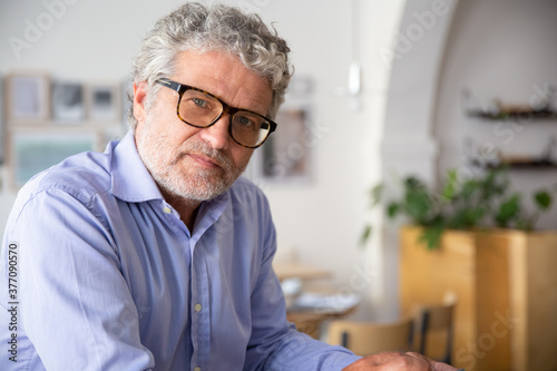 Serious pensive mature business man wearing shirt and glasses, sitting in office cafe, looking at camera. Medium shot, copy space. Business portrait concept