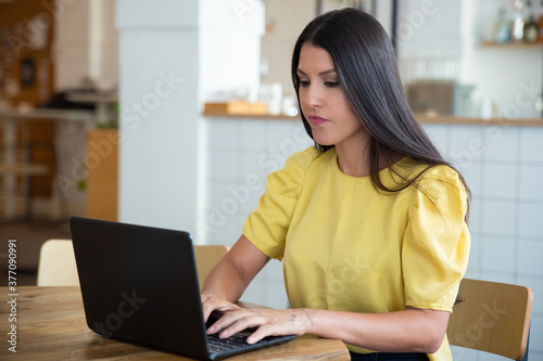 Focused beautiful black haired woman sitting at table in co-working space, using laptop, looking at display and typing. Medium shot. Wireless communication or workplace concept