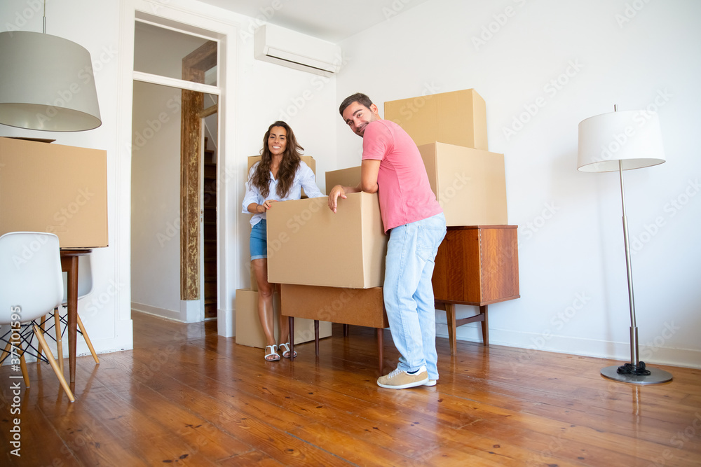 Young couple moving into new flat, leaning on cardboard boxes and furniture indoors, looking at camera and smiling. Full length. New home or property buying concept Young couple moving into new flat, leaning on cardboard boxes and furniture indoors, looking at camera and smiling. Full length. New home or property buying concept