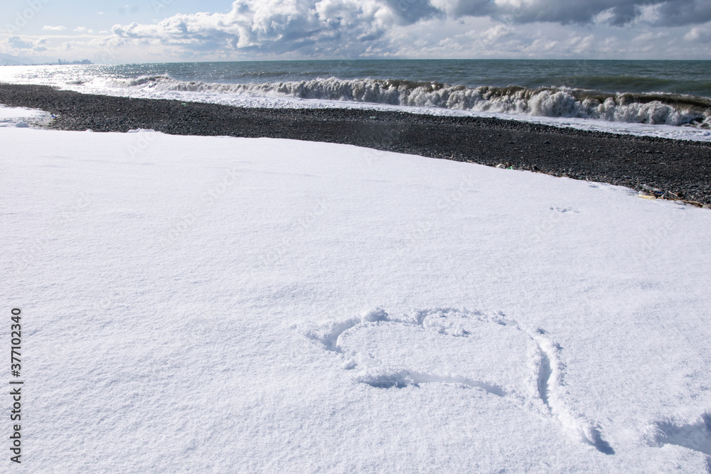 Heart shaped form on white snow with rocky beacj and sea view onthe background. Horizontal blank space image.