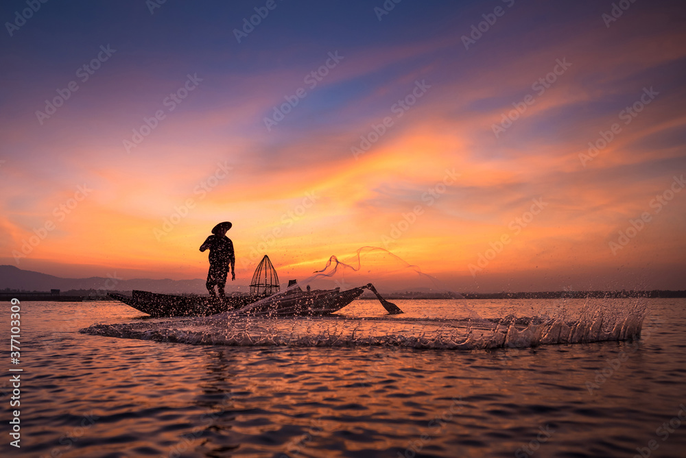 Asian fisherman on wooden boat throwing a net for catching freshwater fish  in nature river in the early morning before sunrise Stock Photo