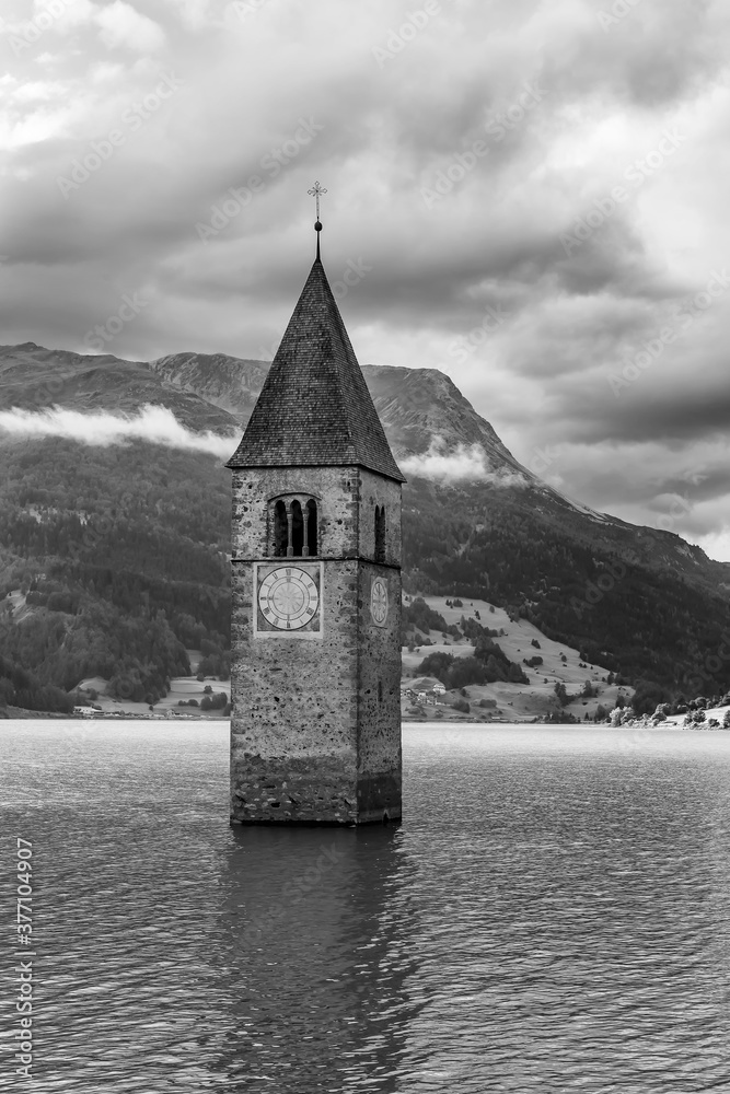 The old bell tower of the church of Curon Venosta submerged in Lake Resia, South Tyrol, Italy, in black and white against a dramatic sky