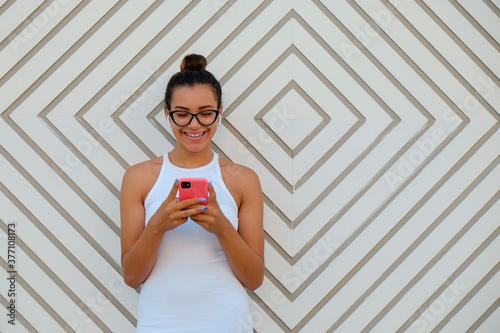 Young beautiful woman of Arabic ethnicity wearing tight white dress holding her smartphone, standing near the wall with abstract geometrical patterns. Close up, copy space, background. photo