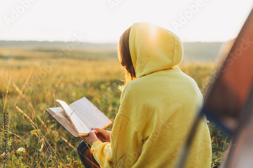 Young woman traveler in yellow hoodie reading book against the tent. Local tourism, weekend trip