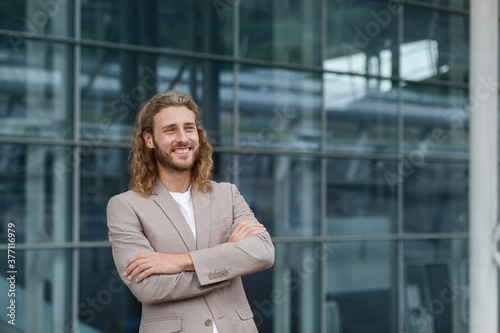 Business portrait of handsome curly smiling man with long hair in casual wear, standing on city street near business center. Confident businessman became successful.