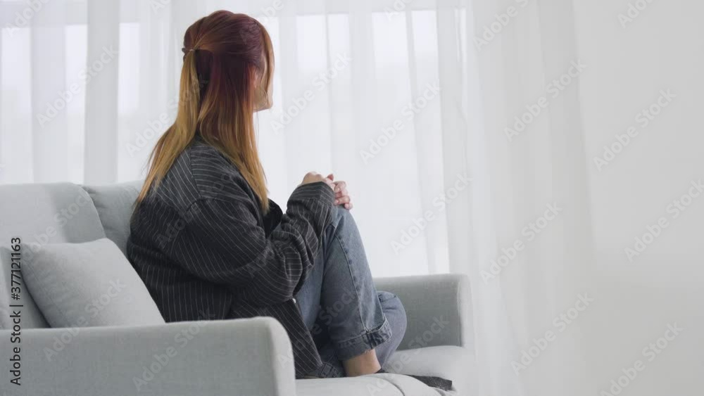 Side view of depressed red-haired young woman sitting on couch and looking out the window. Sad Caucasian lady spending lonely day at home. Heartbreak concept. Side view of depressed red-haired young woman sitting on couch and looking out the window. Sad Caucasian lady spending lonely day at home. Heartbreak concept.