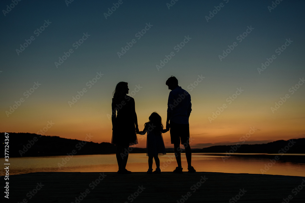 A family of three is silhouetted. Parents hold their daughter's hands.