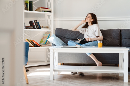 Image of smiling beautiful girl reading book while sitting on sofa