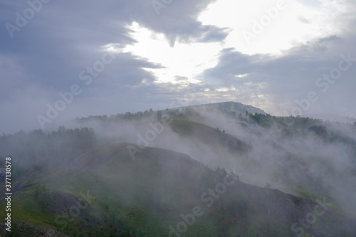 Mountain at sunrise. Fog rises along mountain hollows. Green grass and trees. Small house, dirt road to the distance. Bright sun rays through dense clouds in the sky.